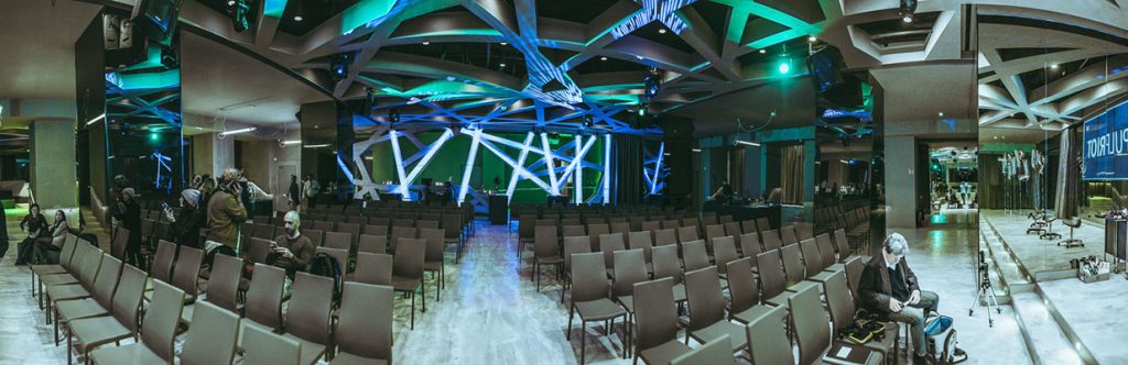Audience seating in a modern venue with a blue-lit stage and sculptural ceiling, rows of empty chairs awaiting a presentation.