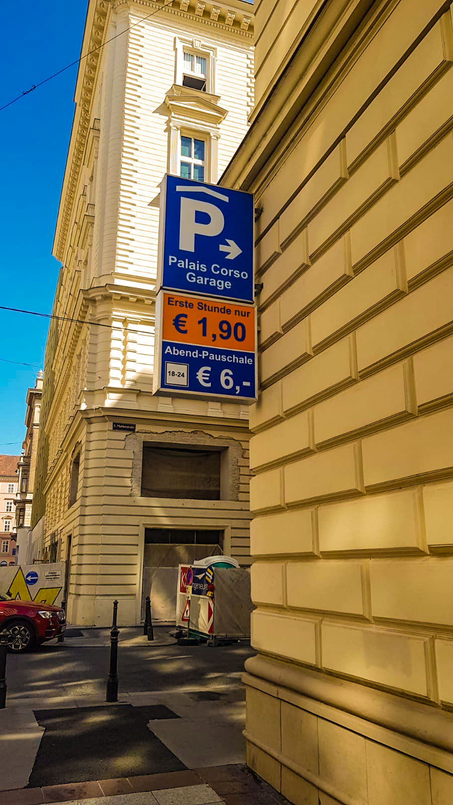 Screenshot - Palais Wertheim Eventlocation Wien Blue parking sign on a beige corner building reads 'Palais Corso Garage' with rate panels: first hour €1,90 and evening flat rate €6,- on a sunny street.