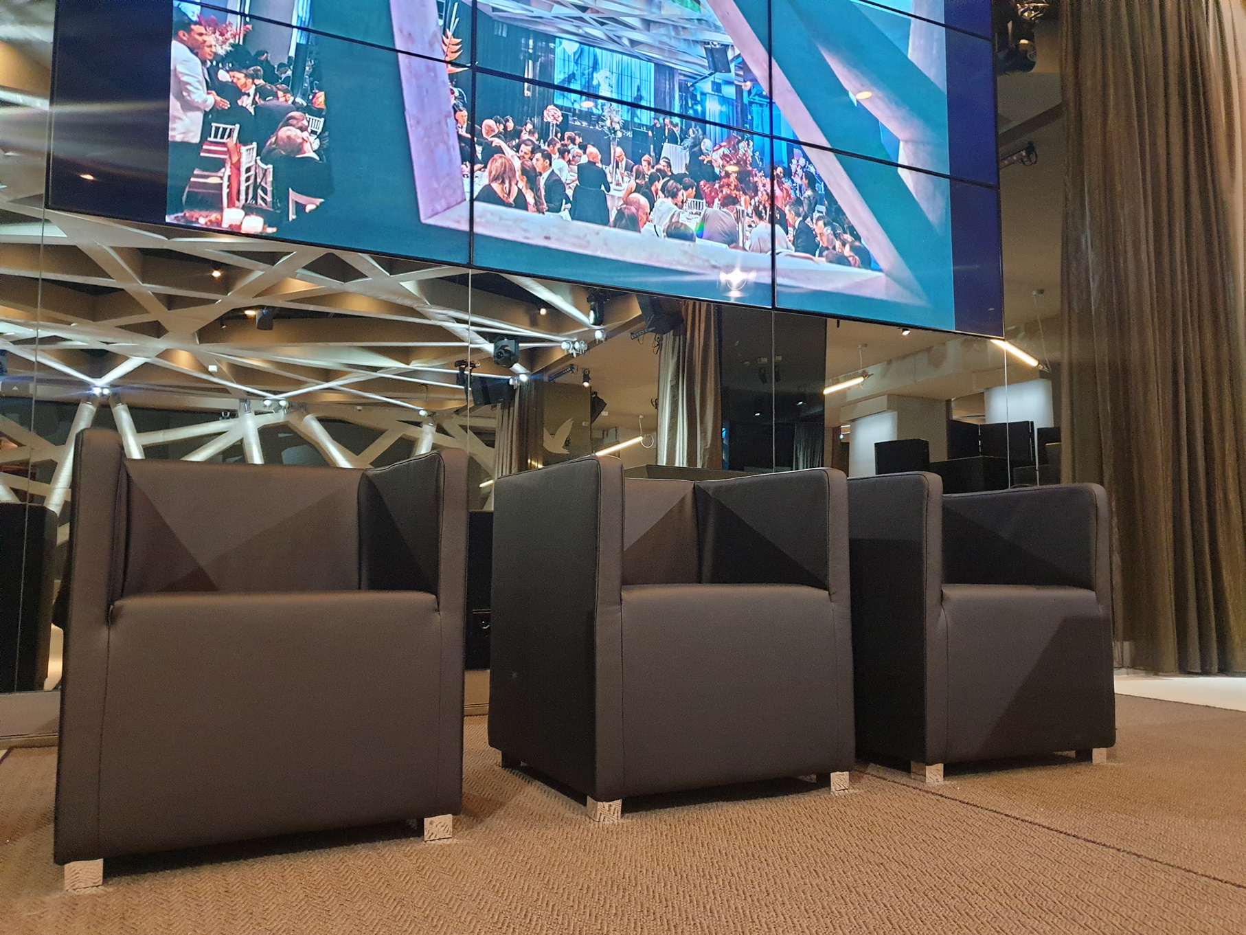 Four black armchairs in a modern lounge facing a large multi-screen video wall showing a crowded event.
