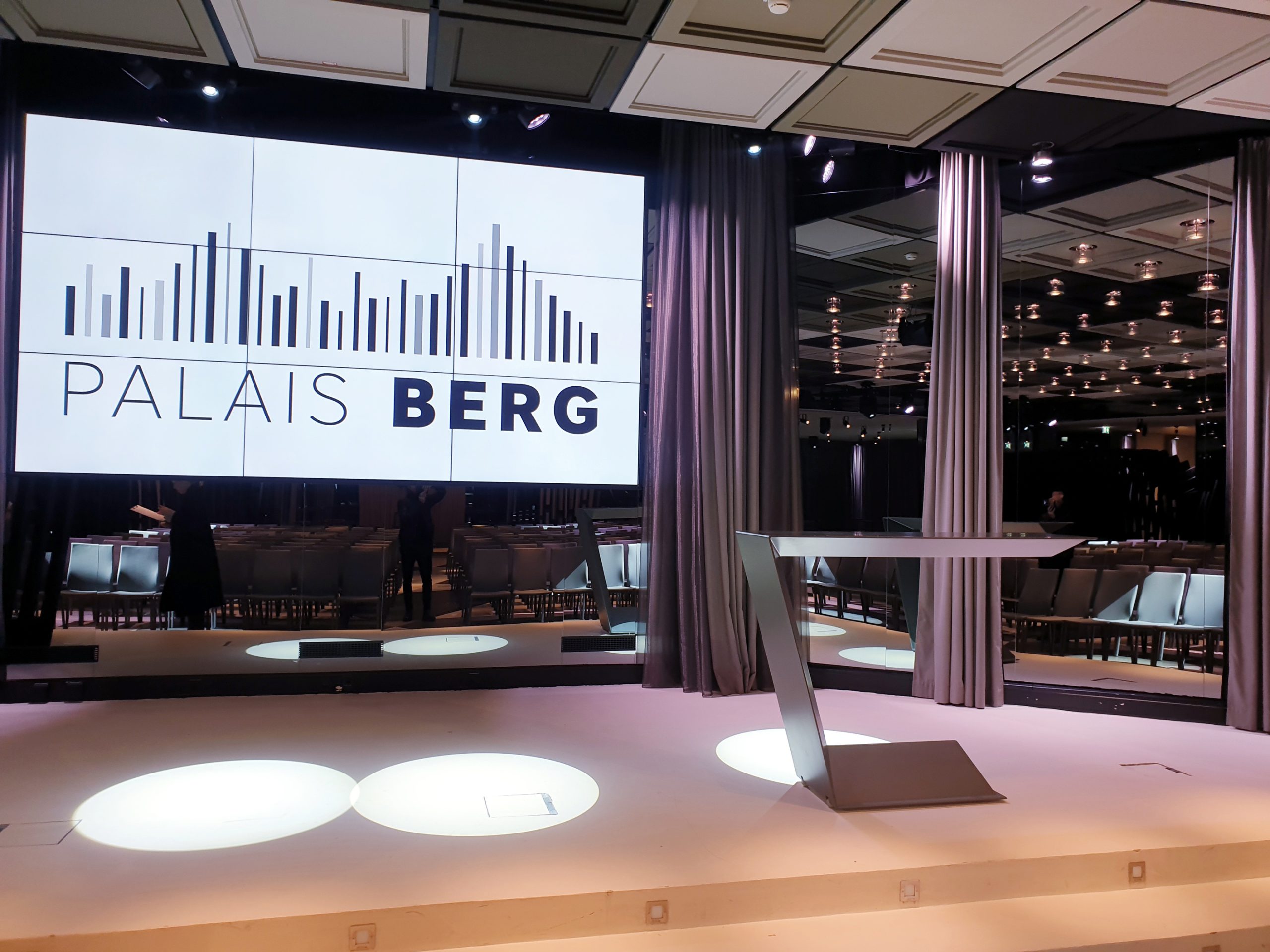 Stage with a modern lectern and a large screen reading PALAIS BERG beside a bar-chart graphic.