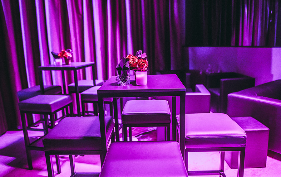 Dimly lit lounge with square tables, purple lighting, and cushioned stools; small flower arrangements and candles on tables.