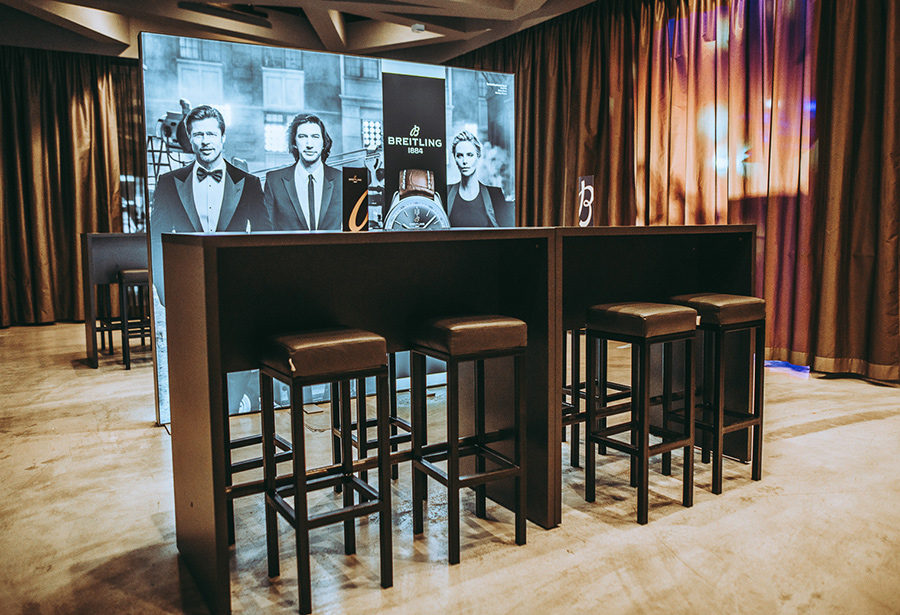 Breitling promotional booth with black high tables and stools, backdrop featuring brand imagery and watches in a curtained event space