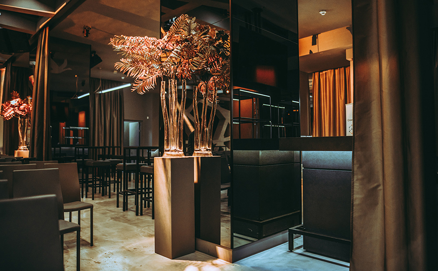 Upscale lounge interior with warm lighting, tall glass vases with ferns on pedestals, and dark seating.